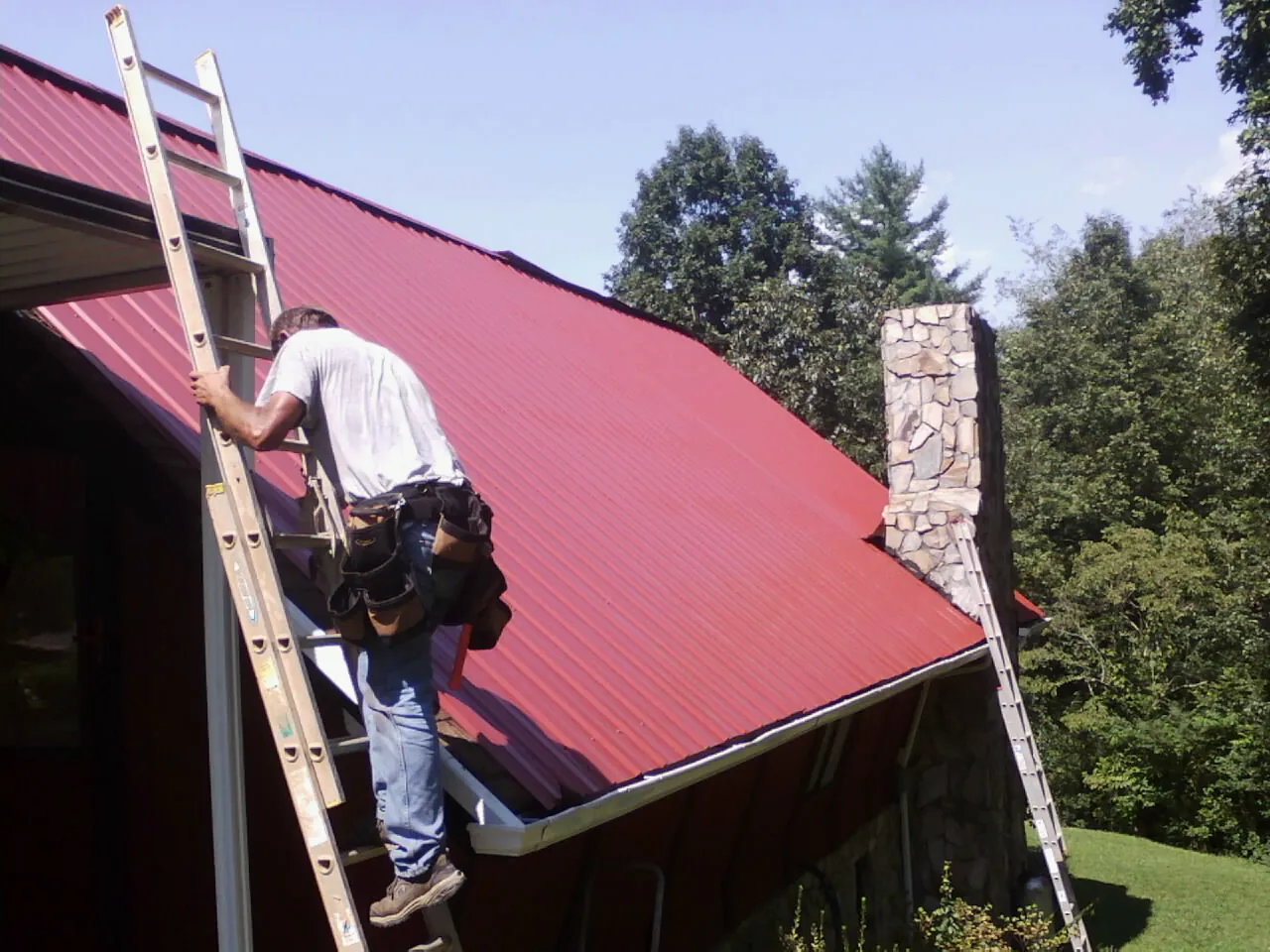 Roofer on a ladder beside a red metal roof and stone chimney — Soffit & Fascia Repair by Roofing Brothers in Fort Montgomery